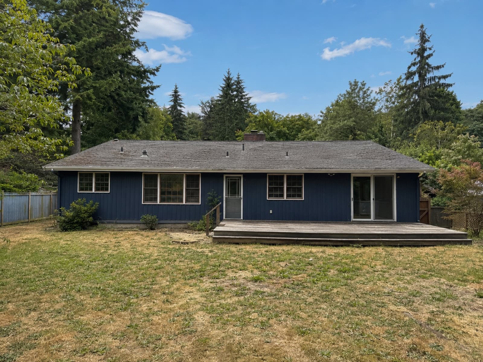 A single-storey ranch home with dark blue siding, a weathered deck, and a patchy dry lawn.